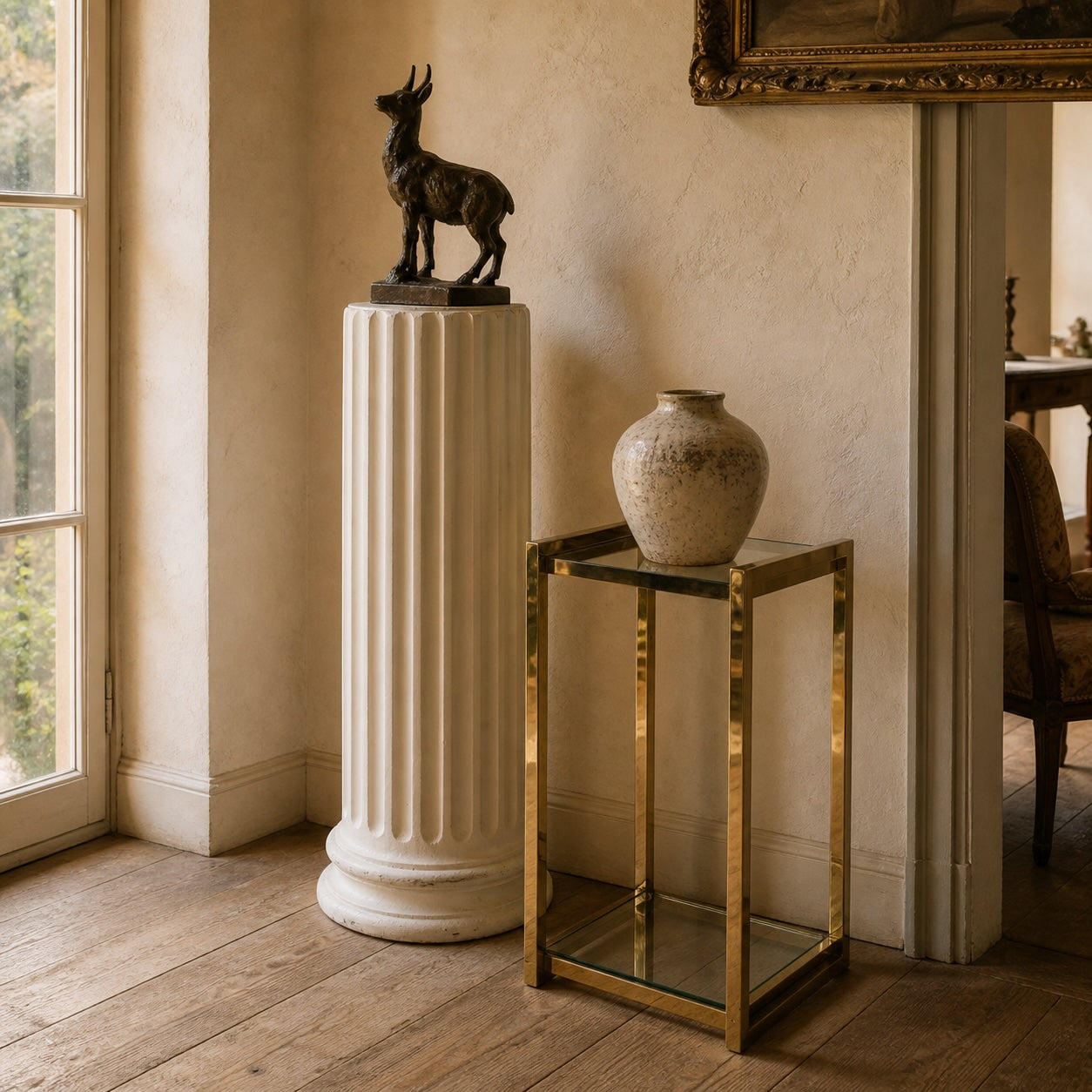 Sunlit French interior corner with fluted white pedestal holding a bronze goat sculpture, beside a brass and glass side table