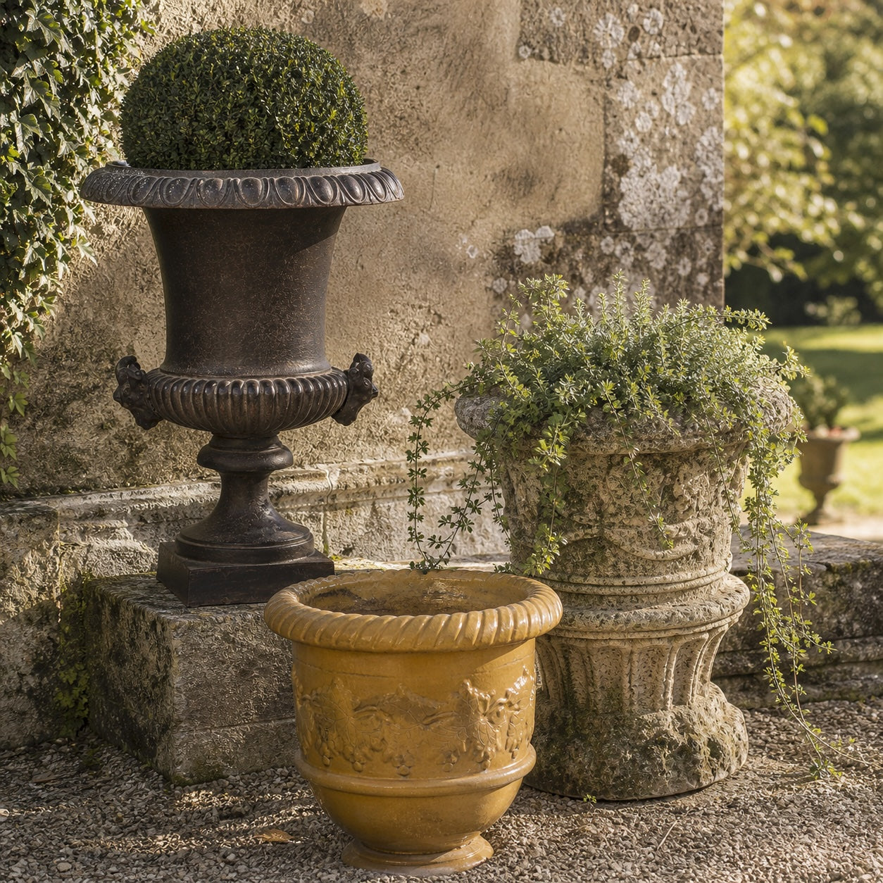 Antique French garden planters on stone steps, including cast iron Medici urn with clipped box topiary, weathered stone planter with trailing rosemary, and mustard glazed terracotta Anduze urn