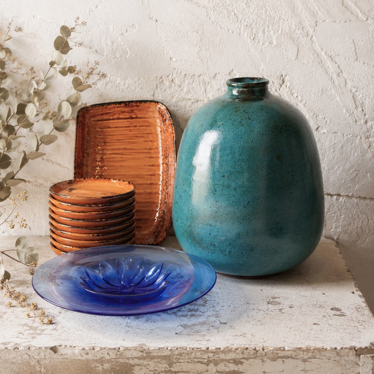 Mediterranean beach house vignette with an aqua glazed French stoneware vase, cobalt blue coupe glass bowl, and terracotta Vallauris plates against a whitewashed plaster wall
